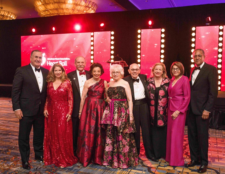Group of nine men and women in formal wear in front of a screen with the American Heart Association Heart Ball Logo.